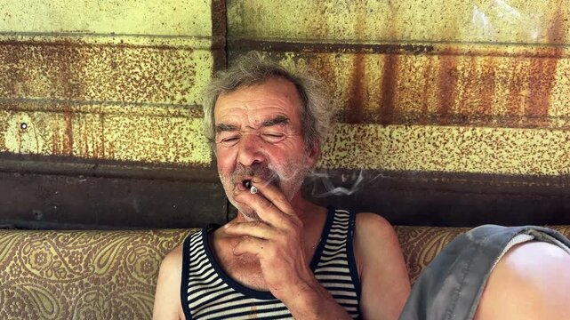 Portrait of homeless adult man sitting on worn couch placed and smoking outdoor. The setting underscores neglect and impermanence, presenting a raw and unfiltered glimpse of difficult living situation