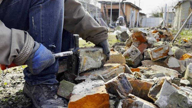 Male hands in protective gloves uses hammer to remove old cement from used bricks during renovation work. The process involves carefully cleaning brick surface to prepare it for reuse in construction