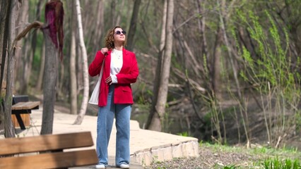 Fototapeta premium A young woman with curly hair in a red jacket walks through a spring park, resting from a working day.