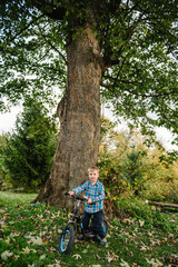 Fototapeta premium Child riding balance bike on a grassy path in the park in autumn. Smiling child having fun outdoors, with colorful trees and fallen leaves in the background.