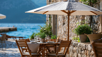 Obraz premium Outdoor dining at a rustic stone cafe terrace shaded by a large sun umbrella in a sunny Mediterranean setting, seaside travel concept, with copy space