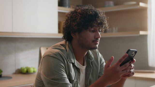 Unhappy Indian Arabian man smartphone receiving bad news at home kitchen upset ethnic male holding mobile phone reading negative message stressed disappointed shocked problem frustrated guy indoors