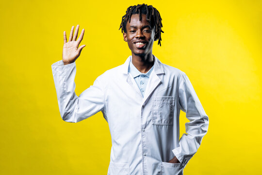 Smiling african man wearing white lab coat, waving hand to camera, standing on yellow background. Doctor concept