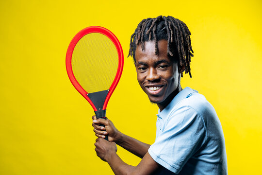 Happy young man with dreadlocks smiling, holding a red tennis racket, on a bright yellow background. Enjoying sports