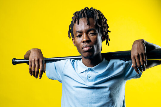 Young black man with dreadlocks holding a baseball bat, projecting a serious and confident attitude on yellow background