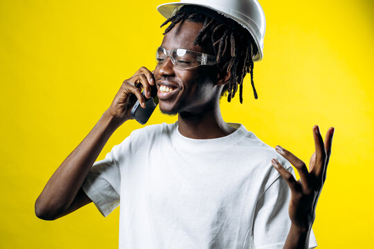 African american construction worker wearing a white hard hat and safety glasses, smiling while talking on a mobile phone
