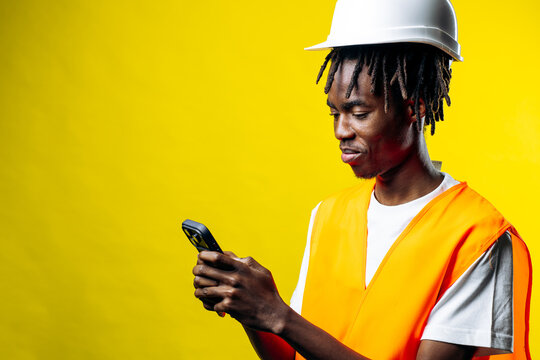 Young man in hard hat and safety vest smiling, actively using smartphone with a yellow background