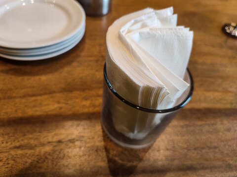 Neat napkins sit in a glass on a wooden table. Stacked plates and a metal cup nearby. Warm lighting enhances cozy restaurant feel. Image captures simple, inviting dining moment