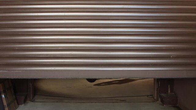 Vintage blue motorcycle standing ready inside a garage with an opening aluminum roller shutter door, creating a strong contrast of light and shadow on the concrete floor