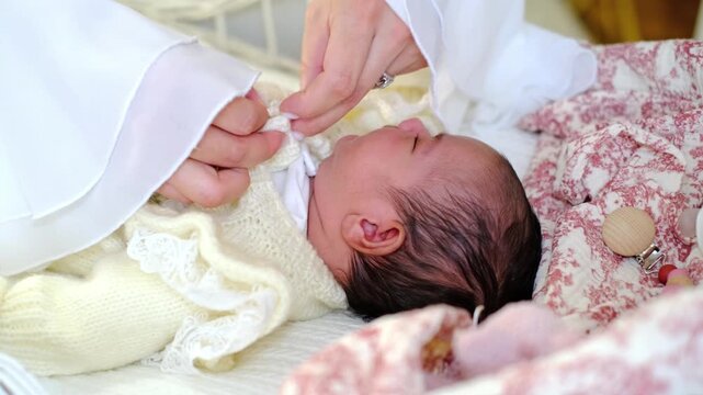The camera lingers over a newborn as adult hands straighten a handmade cream cardigan, using a soft side angle and quiet pace. This scene fits early care, tenderness, craft clothing, and home life.