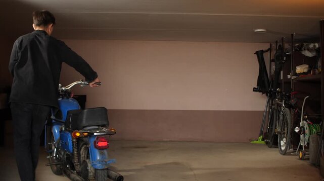 Young man pushing an old motorcycle out of storage in a dimly lit garage, showing hobby, transportation, and retro lifestyle