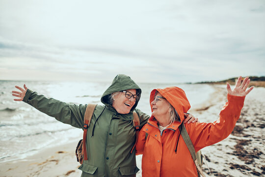 Two senior women embracing on windy beach in raincoats