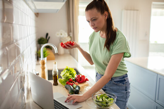 Woman preparing salad with fresh vegetables while using laptop in home kitchen