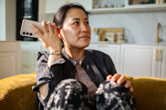 Thoughtful woman listening to voice message on smartphone in modern kitchen