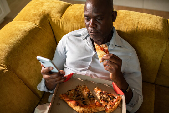 Man eating pizza and using smartphone on sofa at home