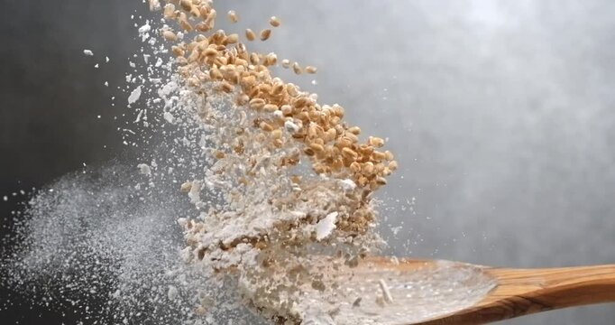 Wheat grains and flour falling from spoon creating powder cloud in slow motion