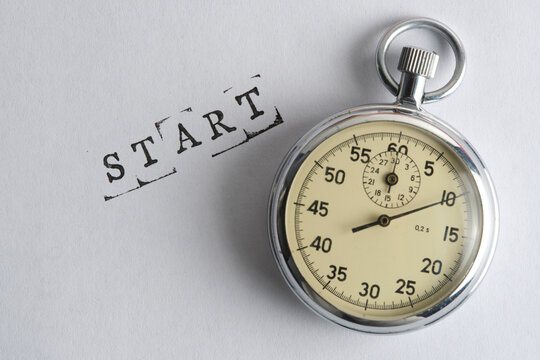 Classic mechanical stopwatch resting next to word START printed on light textured paper background.