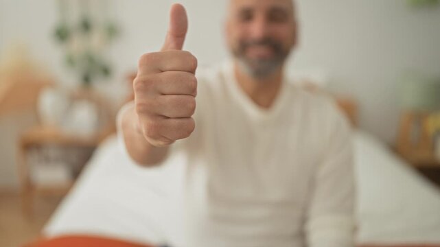 Man seated on bed in a building extends his thumb toward camera showing a clear thumbs down gesture and visible hand detail; disapproval.