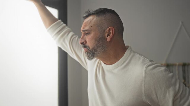 Man with arm raised and hand on window frame, eyes closed, leaning in studio wearing cream sweater; quiet contemplation.