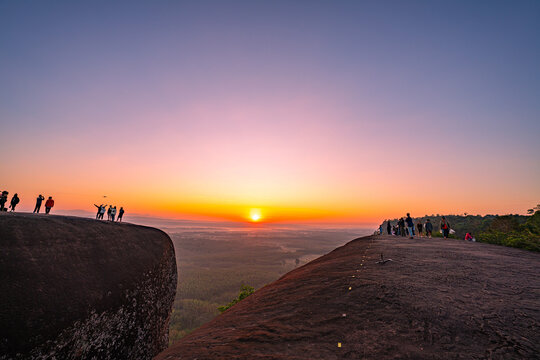 Epic sunrise at a famous Thai viewpoint, capturing tiny human figures on a colossal rock ledge above a sea of trees, evoking adventure, scale, and serenity.