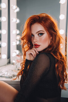 Elegant redhaired woman posing in front of illuminated vanity mirror with glamorous makeup, soft lighting, and sophisticated beauty portrait in modern dressing room