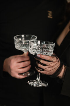 Cheers. Close-up of a man and a woman in black clothes holding elegant crystal coupe glasses. They are making a toast. Dark moody atmosphere, sophisticated celebration, evening party concept.