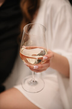 Closeup of a woman's hand holding a glass of white wine. She wears a white silk robe and has a french manicure. The setting is indoors, during a wedding morning.