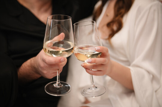 A romantic close-up of a man's and a woman's hands clinking glasses of white wine. The setting is indoors, during a wedding morning or celebration. Soft focus background.
