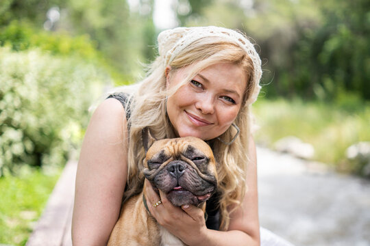 Happy young woman hugging adorable French bulldog while relaxing outdoors in scenic park with warm natural lighting
