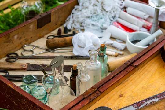 Vintage wooden medical chest filled with historical surgical tools, glass bottles, cupping glasses, bandages, mortar, pestle, scissors, and a bone saw arranged in natural daylight.