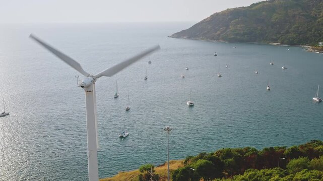 Wind turbine spinning above Phukets green coastline and calm bay, generating clean renewable energy as anchored sailboats rest in tranquil tropical waters during daytime. Aerial static shot