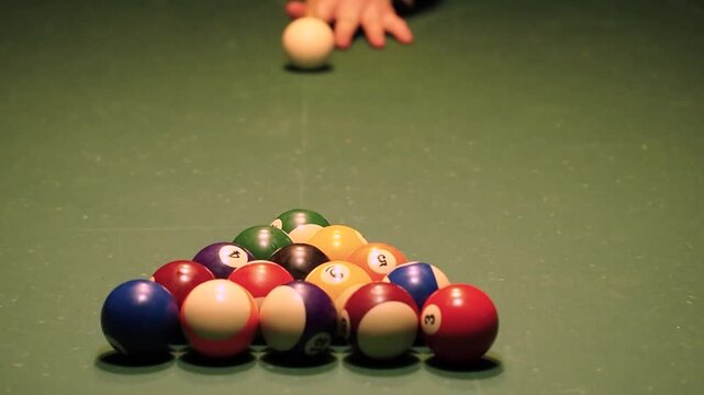 High angle view of a billiard table with a racked set of numbered balls and a white cue ball. A hand positions the cue ball before a cue stick strikes it, causing the balls to scatter on felt. A game