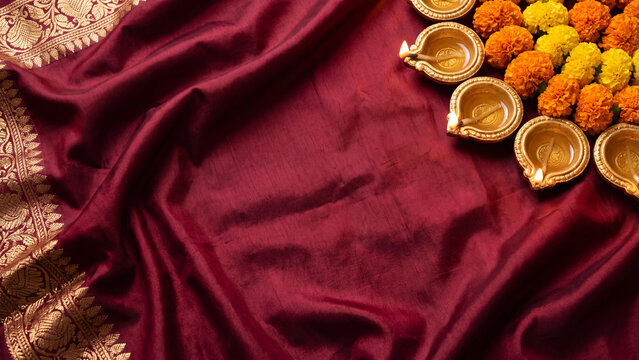 Maroon silk fabric with golden woven border styled with brass oil lamps and marigold flowers with copy space for Diwali greeting cards invitations cultural marketing and elegant background design