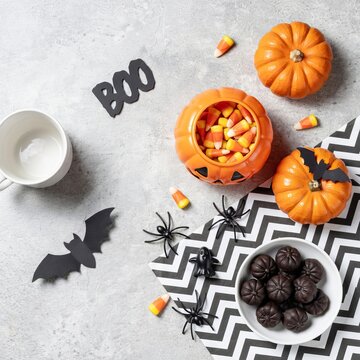 Halloween flat lay featuring a pumpkin candy bowl filled with candy corn, black paper bats, plastic spiders, and a bowl of dark chocolate truffles on a textured gray surface with chevron paper