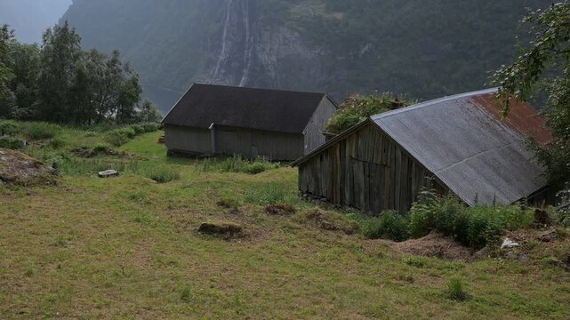 Weathered wooden barns on a grassy hillside with fjord mountains and waterfall in the background.