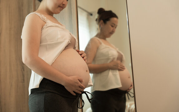 Close-up of a pregnant woman in a white lace top holding her belly with both hands, her reflection visible in a mirror behind.