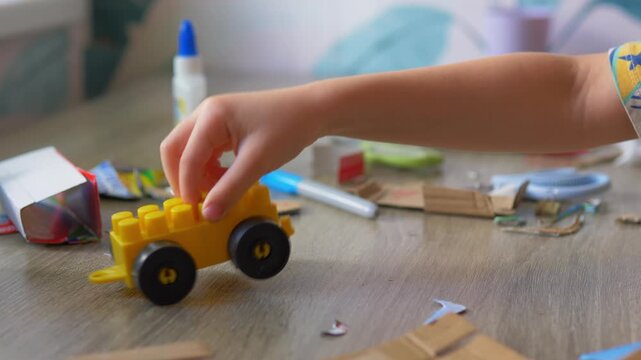 Child's hand holding a yellow block toy car playing above a messy desk with craft supplies