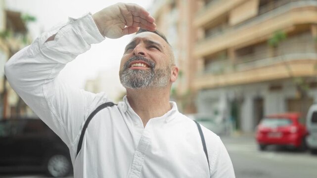Man shields eyes with hand on sunlit street, wearing white shirt and backpack strap while smiling and looking up; hopeful curiosity.