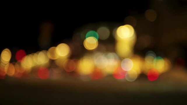 Static wide angle view of urban street at night with heavy bokeh effect. Out of focus city lights appear as circular orbs in yellow red and green. Constant light movement against black background