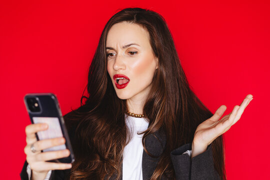 Shocked confused young woman looking at smartphone screen with open mouth gesturing in outrage wearing black blazer with red lipstick, isolated on solid bright red background expressive stock portrait