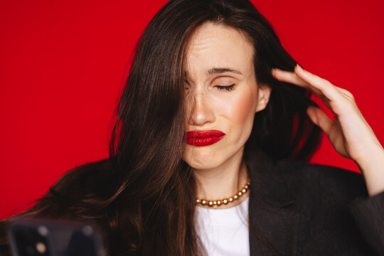 Distressed sad young woman closed eyes hand on head facepalm pouting lips reading terrible news text message on smartphone wearing dark blazer red lipstick, isolated on studio bright red background