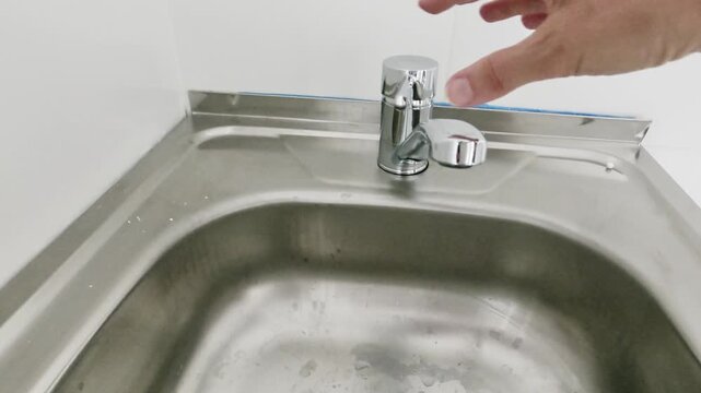 A first-person perspective view of a stainless steel sink and chrome faucet on a white cabinet. A hand lifts the lever to start water flow and pushes it down to stop the flow in a bright tiled room