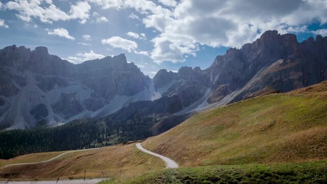 A winding path through a grassy valley leading towards majestic, rugged mountains under a partly cloudy sky.