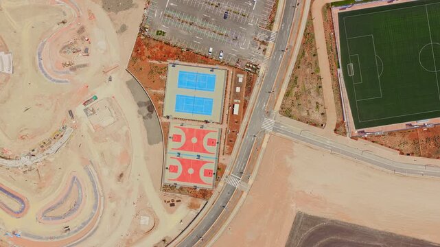 Aerial zenith view slowly spiraling over a new residential area, featuring basketball courts, tennis courts, a soccer field, and an adjacent construction site for urban development
