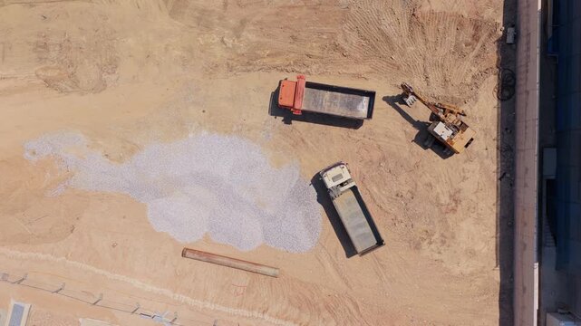Top-down aerial view of a dumper truck unloading a pile of gravel at a construction site, with an excavator nearby, showcasing the initial stages of building a new foundation