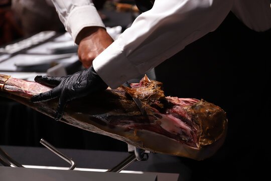 experto del jam&oacute;n ib&eacute;rico manipulando con sus manos con guantes y u cuchillo jamonero para limpiar la costra externa del jam&oacute;n