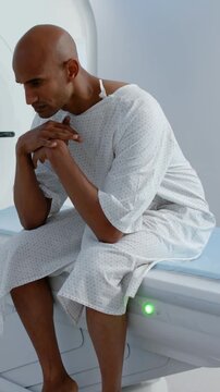 Vertical video: Sitting man in gown fidgeting hands and lifting gaze at scan bed preparing for scan