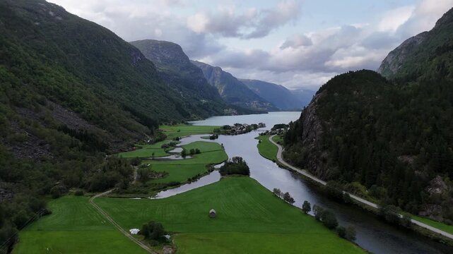 A beautiful aerial view of a winding river flowing through a lush green valley towards a large lake surrounded by majestic mountains under a cloudy sky.