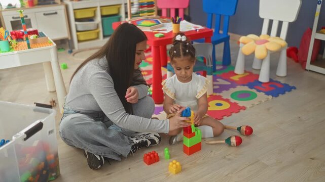 Woman stacking colorful blocks with little girl on foam playmat inside a building; playful learning.