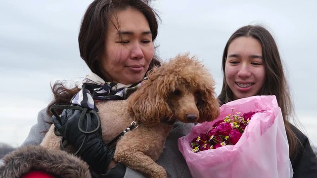 A woman holding a curly poodle, a teenage girl holding a pink bouquet of flowers and her young son on a calm beach, under a soft overcast sky with a view of the coast in the distance, a casual